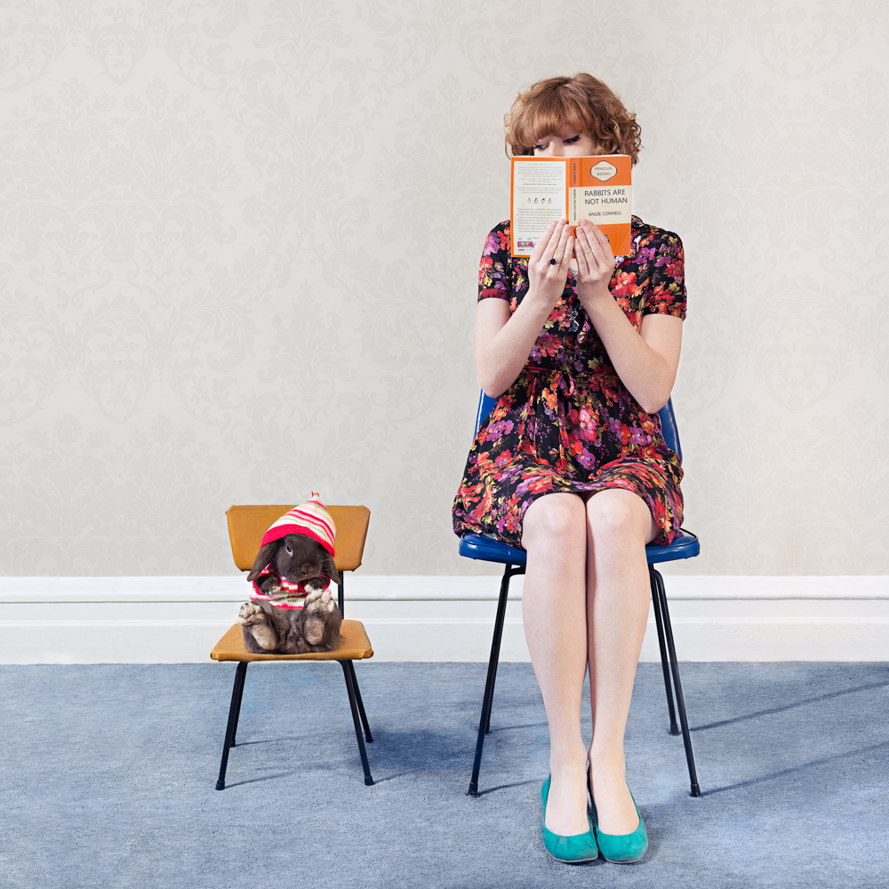 girl sitting on a chair reading a book with her rabbit sitting next to her on a chair