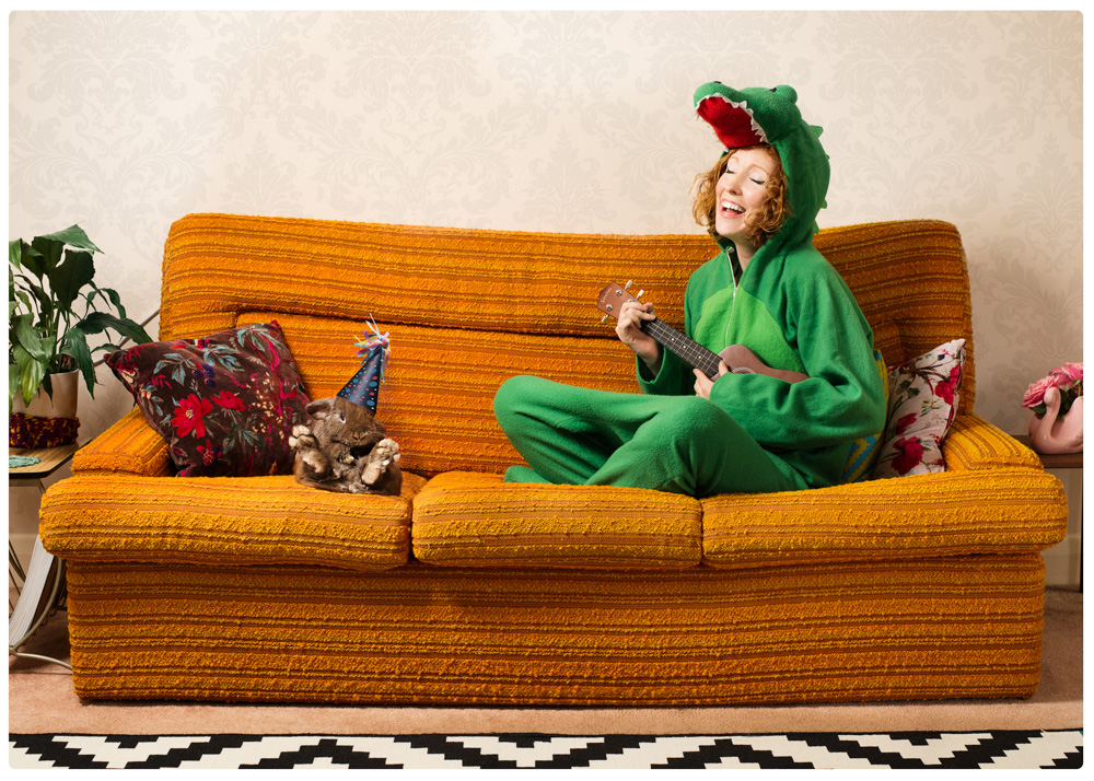 girl dressed in croc onesie playing a ukulele on the couch with her rabbit